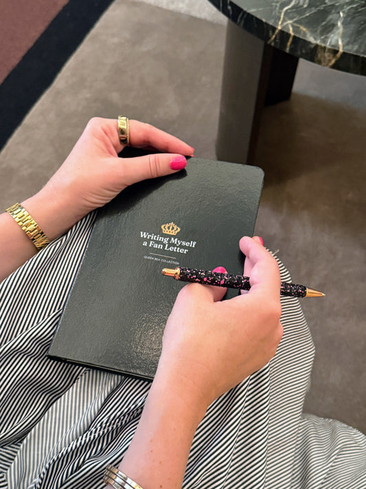 A hand holding a gold pen over the black Writing Myself a Fan Letter journal