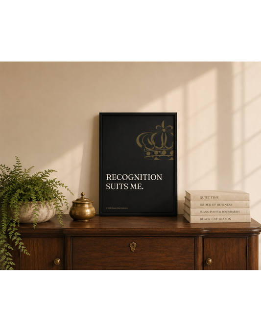 Recognition Suits Me canvas print in a black floating frame, propped on a wood console with a fern, brass vessels, and a stack of books