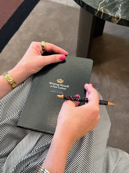 A hand holding a gold pen over the black Writing Myself a Fan Letter journal