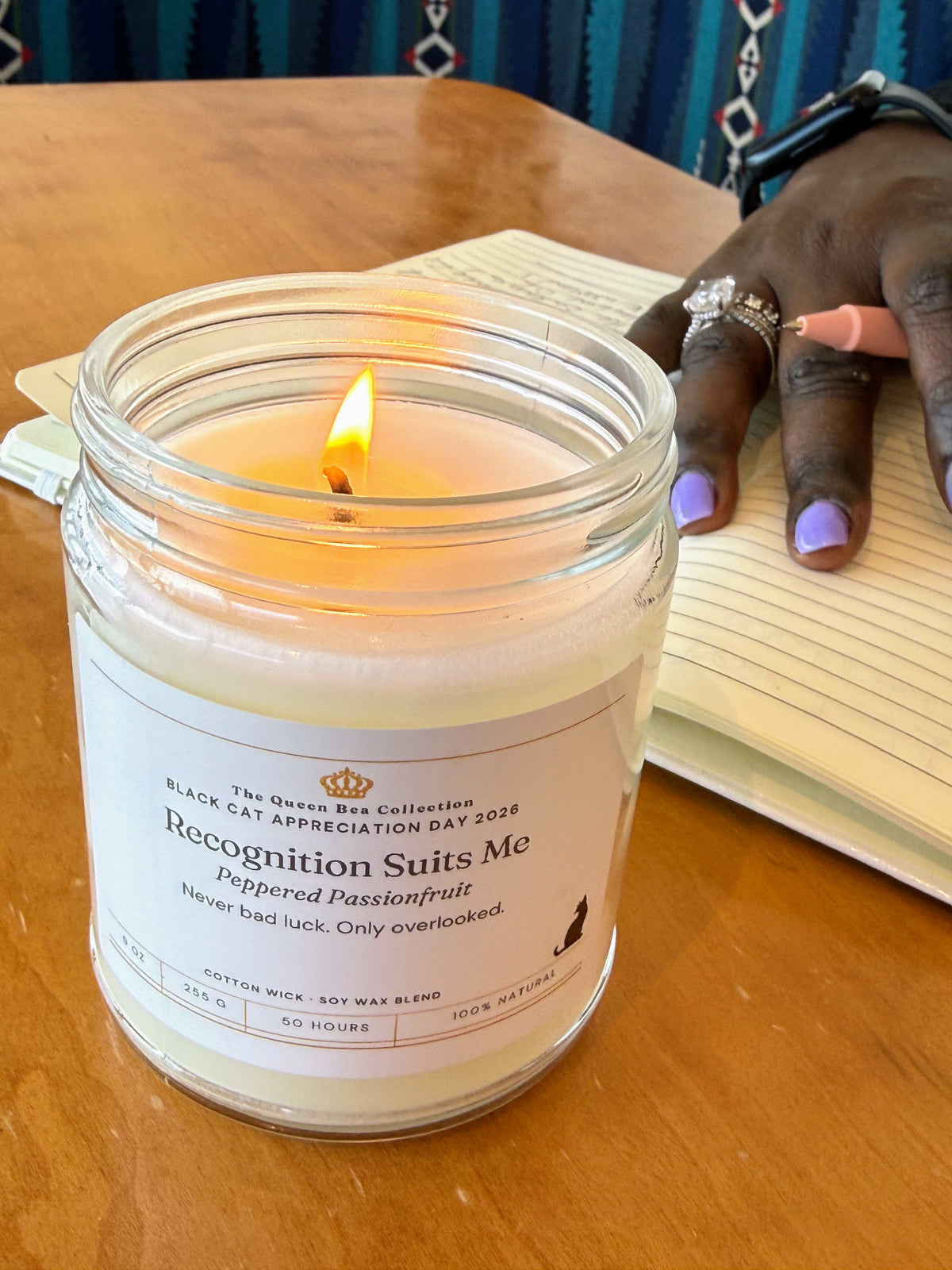 A hand writing in a journal beside the lit Recognition Suits Me candle