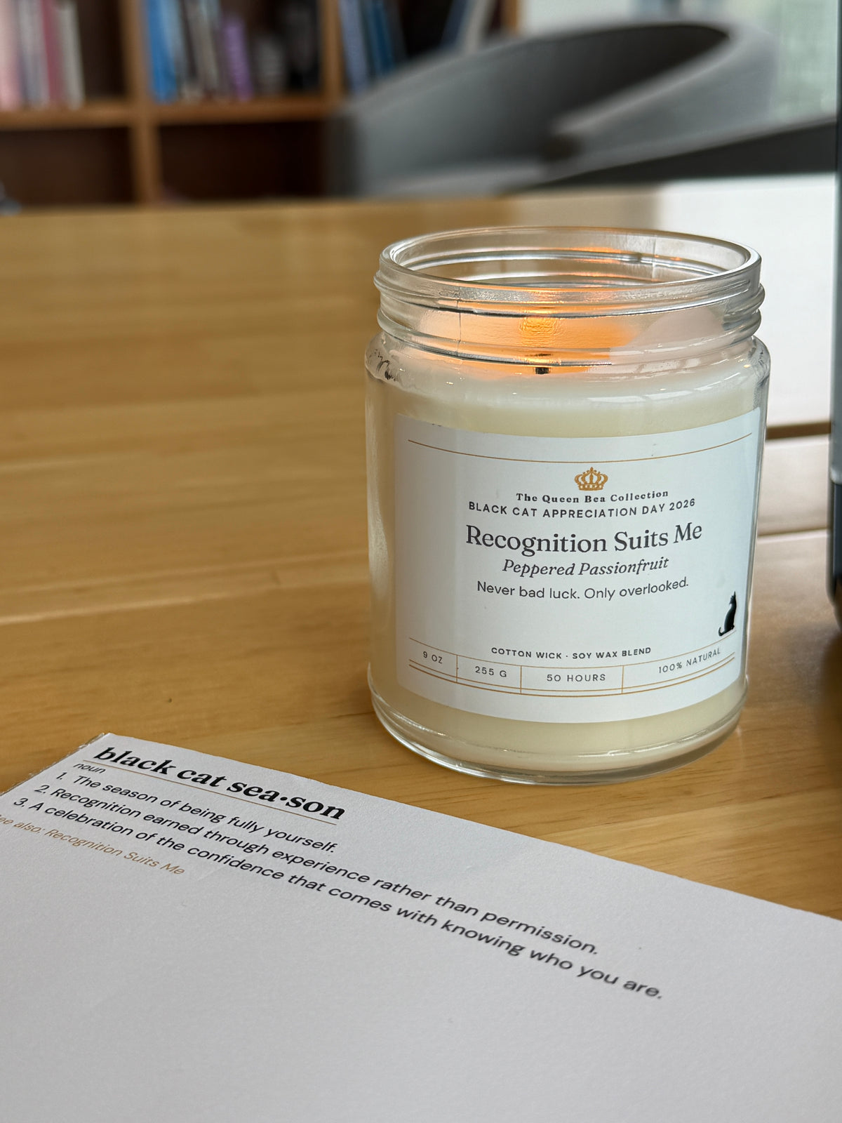 Recognition Suits Me candle and the Black Cat Season notepad on a desk, bookcase behind