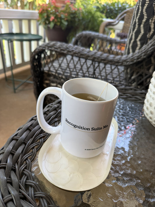 Recognition Suits Me mug with tea on a porch table, wicker chairs and flowers behind